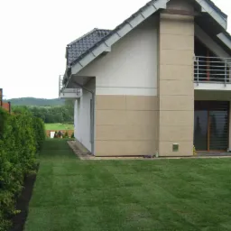 Nowoczesny dom z zadbanym trawnikiem i szpalerem tuj, widok z perspektywy ogrodu. W tle zieleń drzew i pagórki. Widoczne dwie osoby pracujące w oddali.