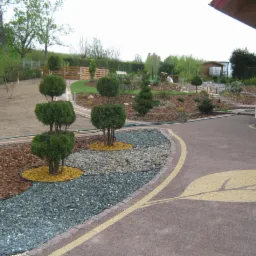 Aranżacja ogrodu z ozdobnymi drzewkami formowanymi, ścieżką z kamienia i podjazdem z kostki brukowej z motywem liścia. Urozmaicone nasadzenia i rabaty.