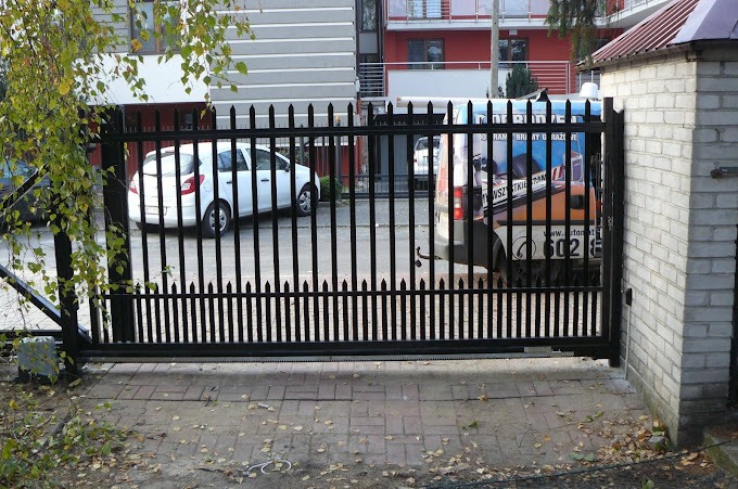 Czarna, metalowa brama przesuwna z ostrymi zakończeniami pionowych elementów, widoczna na tle budynku mieszkalnego i samochodu dostawczego z reklamą firmy montującej automatykę do bram.