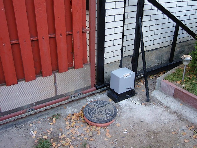 Czerwona, drewniana brama przesuwna z zamontowaną automatyką i zębatką, widoczny szary napęd bramy z modelem 740, studzienka kanalizacyjna obok.