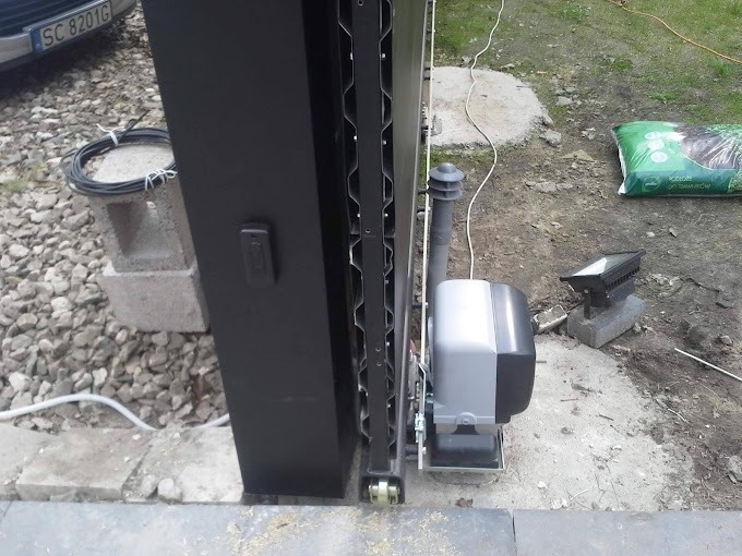 Fragment automatycznej bramy przesuwnej z widocznym napędem, czarną konstrukcją i elementami mocującymi, w tle gruz, trawa i worek ziemi.