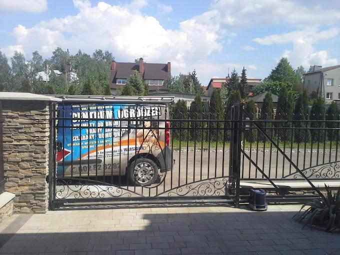 Czarna brama przesuwna z ozdobnymi elementami, widoczny automat do bramy i firmowy van z reklamą w tle, kostka brukowa na podjeździe.
