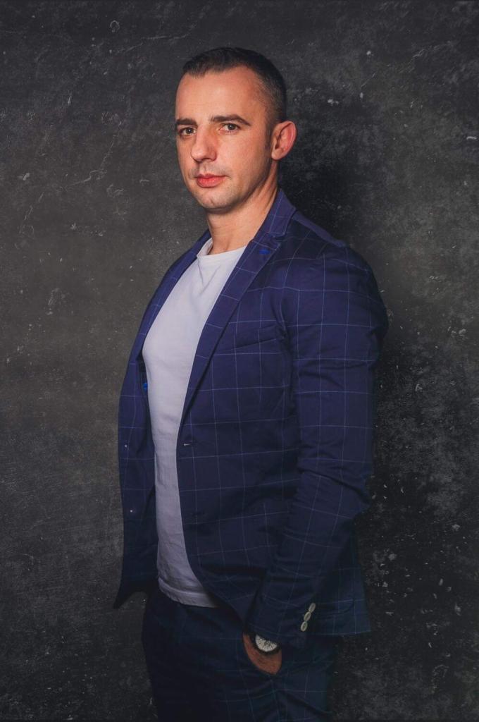 Mężczyzna w niebieskiej marynarce w kratę i białym t-shircie na tle szarej ściany. Portret biznesowy z zegarkiem w kieszeni.