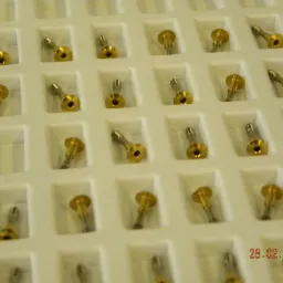 Precyzyjne, złoto-srebrne elementy tokarskie/CNC w białej, kasetowej wytłoczce. Elementy z gwintem i otworem, ułożone równo. Data 28.02.2014 w prawym dolnym rogu.