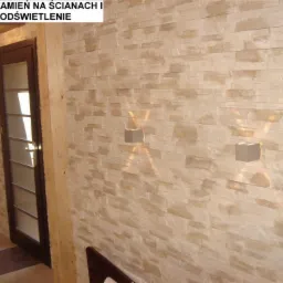 Ściana wyłożona dekoracyjnym kamieniem w odcieniach beżu, oświetlona dwoma kwadratowymi kinkietami, widoczna w Częstochowie, z ciemną stolarką drzwiową w tle.