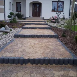 Schody wejściowe do domu wyłożone jasną kostką z szarą obwódką, prowadzące od ogrodu z kamieniami i roślinami do drzwi wejściowych.