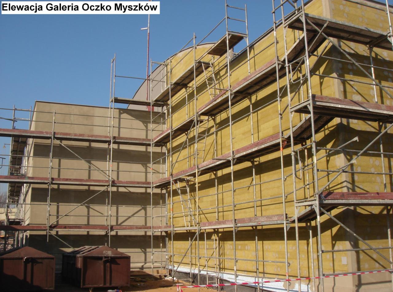 Budynek w trakcie ocieplania, widoczne rusztowania i warstwa izolacyjna w kolorze żółtym, fragment elewacji z nałożonym tynkiem, kontenery na gruz w dolnej części zdjęcia.