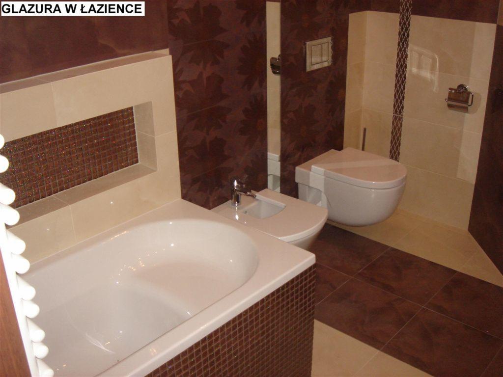 Łazienka z brązowymi i beżowymi płytkami, mozaika nad wanną, wisząca toaleta i bidet, brązowa ściana z motywem liści.