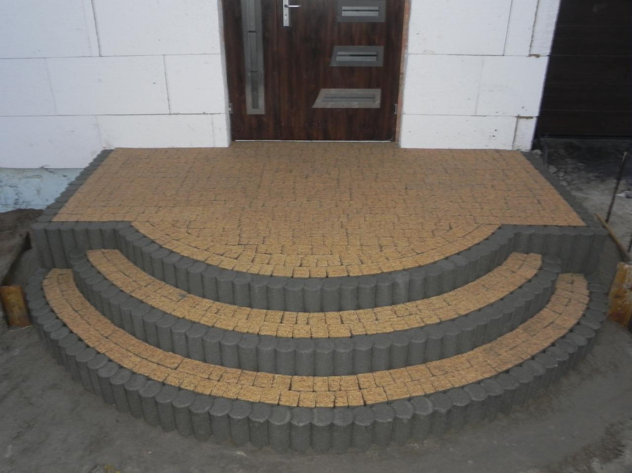 Półokrągłe schody wejściowe z kostki brukowej w odcieniach beżu i szarości, prowadzące do drzwi. Widoczne bloczki betonowe ściany budynku.