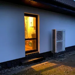 Zewnętrzna jednostka grzewcza na metalowej podstawie przy ścianie domu, widok przez okno na instalację wewnętrzną, wieczorne oświetlenie.