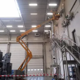 Montaż klimatyzacji w hali przemysłowej z użyciem podnośnika koszowego, widoczne jednostki zewnętrzne na ścianie i rury wentylacyjne.