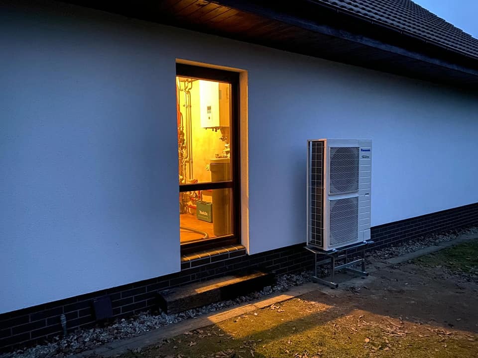 Zewnętrzna jednostka grzewcza na metalowej podstawie przy ścianie domu, widok przez okno na instalację wewnętrzną, wieczorne oświetlenie.