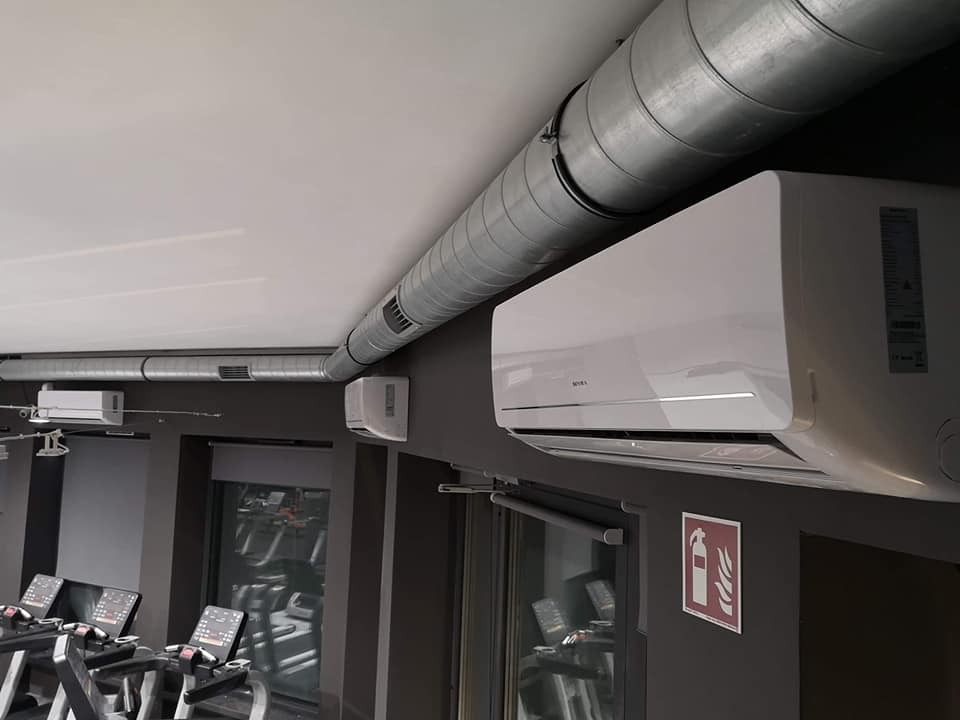 Biała klimatyzacja ścienna zamontowana w siłowni, widoczne przewody wentylacyjne pod sufitem i sprzęt do ćwiczeń w tle.