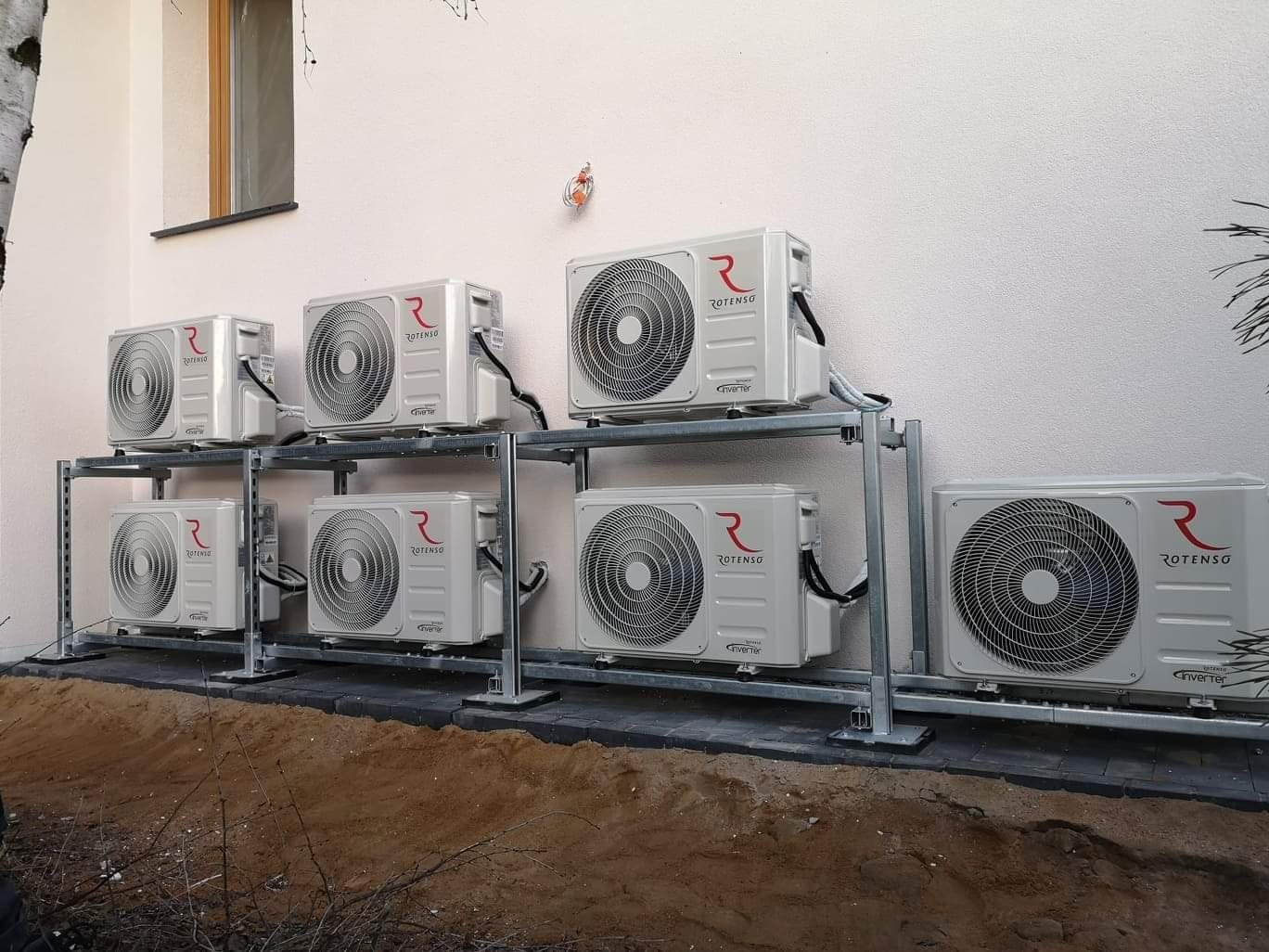 Zewnętrzne jednostki klimatyzacji Rotenso, zamontowane na metalowym stelażu przy ścianie budynku. Widoczne okno i fragment drzewa.