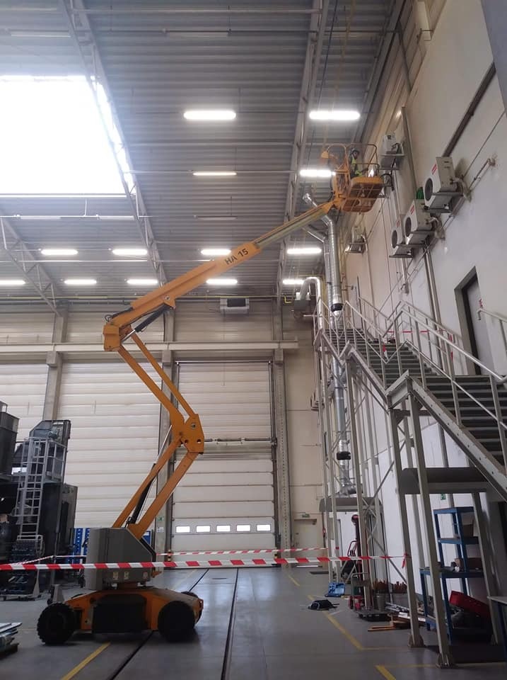 Montaż klimatyzacji w hali przemysłowej z użyciem podnośnika koszowego, widoczne jednostki zewnętrzne na ścianie i rury wentylacyjne.