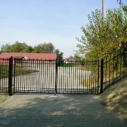 Czarna, metalowa brama dwuskrzydłowa z pionowymi prętami i przyległe ogrodzenie, widok z zewnątrz na brukowany podjazd, w tle budynki z czerwonymi dachami i drzewa.