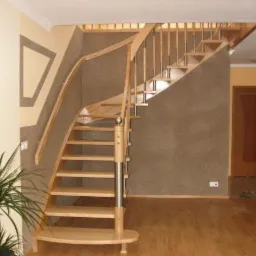 Jasne, drewniane schody z metalowymi elementami balustrady, zakręcające w lewo, w przestronnym wnętrzu z parkietem i rośliną doniczkową.