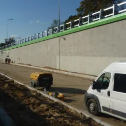 Przygotowanie podłoża pod drogę: widoczna ściana oporowa, maszyna do zagęszczania gruntu i biały samochód dostawczy z logo firmy NIWA.