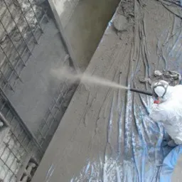 Pracownik w kombinezonie ochronnym nanosi torkret na betonową konstrukcję zbrojoną siatką, zabezpieczoną folią.