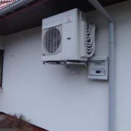 Zewnętrzna jednostka klimatyzacji Mitsubishi zamontowana na białej ścianie domu, obok skrzynka elektryczna i rynna spustowa, widoczne okno z brązową ramą.