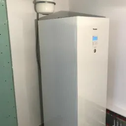 Biała pompa ciepła Panasonic zainstalowana w rogu pomieszczenia z widocznym zbiornikiem wyrównawczym i instalacją grzewczą w podłodze.