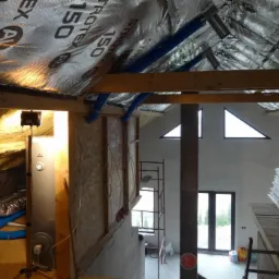 Wnętrze w trakcie instalacji systemu z niebieskimi rurami wentylacyjnymi pod folią izolacyjną dachu, widoczny także rekuperator, surowe ściany i okna trójkątne.