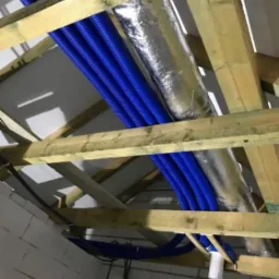 Niebieskie rury wentylacyjne i srebrne przewody izolowane zamontowane pod drewnianą konstrukcją dachu w surowym stanie.