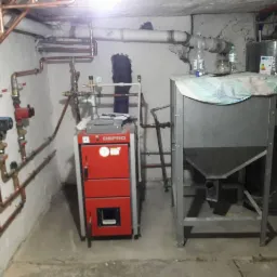 Piwnica z zainstalowanym czerwonym piecem na paliwo stałe marki DEFRO, miedzianymi rurami instalacji grzewczej na ścianie, zbiornikiem na wodę i zasobnikiem na opał z butelkami na górze.