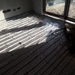 Rury ogrzewania podłogowego rozłożone na izolacji w surowym wnętrzu, widoczne okno i częściowo samoch&oacute;d na zewnątrz.