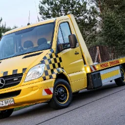 Ż&oacute;łta laweta Mercedes z czarno-białą szachownicą na masce, tablicą rejestracyjną NSZ 88VH i napisem 'Pomoc Drogowa' na tle drzew i drogi.