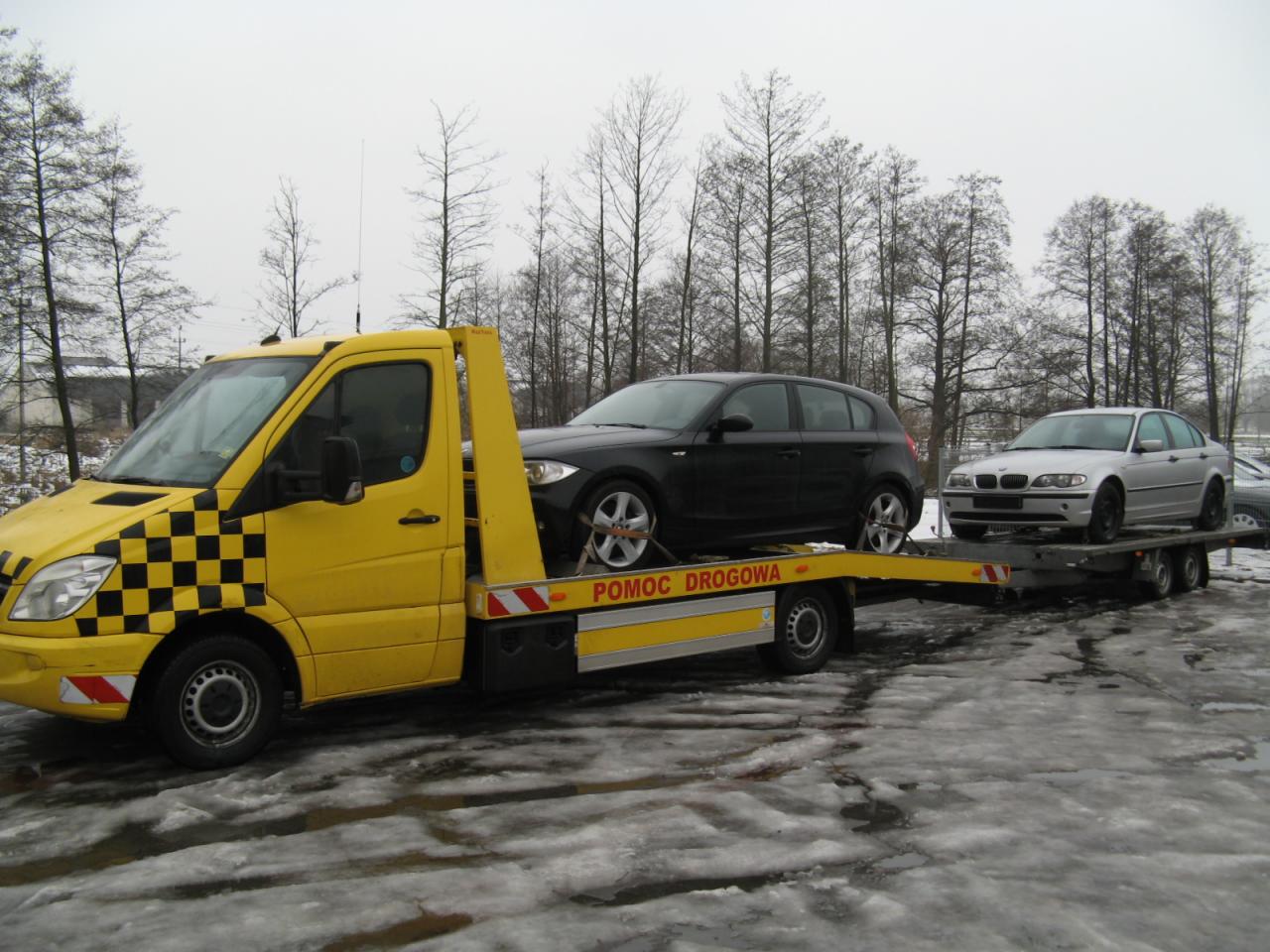 Żółta laweta z szachownicą na kabinie przewozi dwa samochody osobowe na tle zimowego krajobrazu. Widoczny napis 'Pomoc Drogowa'.