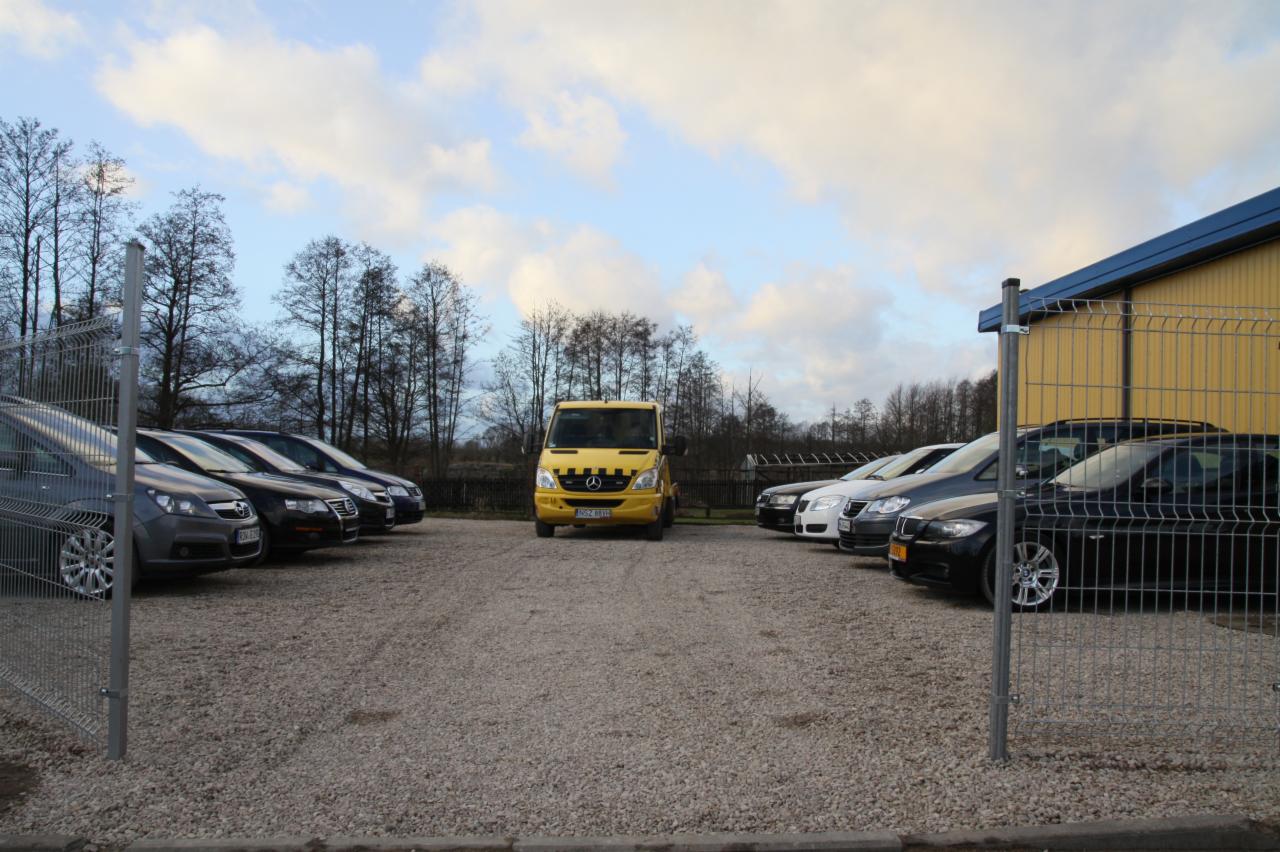 Żółty bus Mercedes-Benz na żwirowym placu, otoczony rzędami samochodów osobowych, widoczne ogrodzenie i budynek w tle. Transport aut, parking.