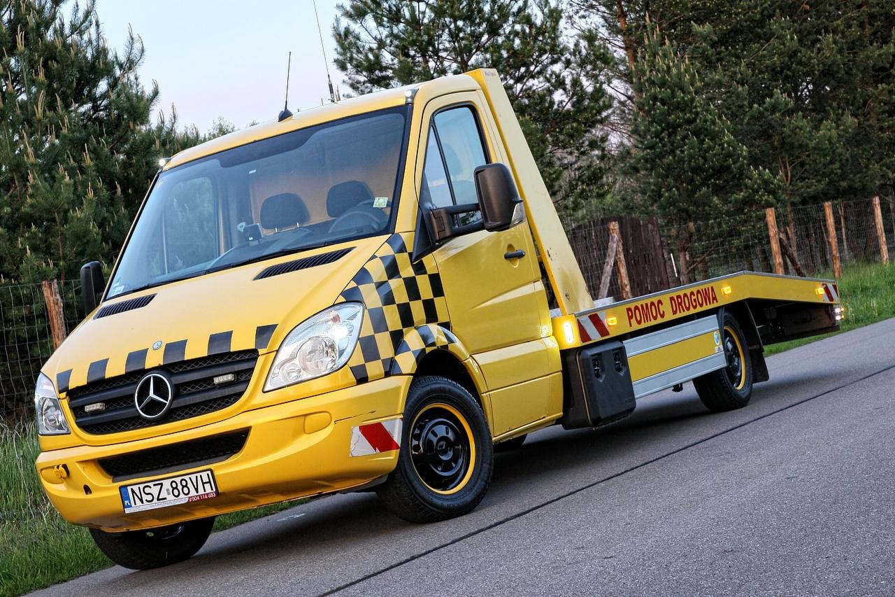 Żółta laweta Mercedes z czarno-białą szachownicą na masce, tablicą rejestracyjną NSZ 88VH i napisem 'Pomoc Drogowa' na tle drzew i drogi.