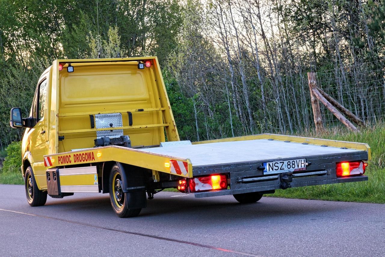 Żółta laweta z napisem 'Pomoc Drogowa' widziana z tyłu na drodze, z lasem w tle i widoczną tablicą rejestracyjną NSZ 88VH.