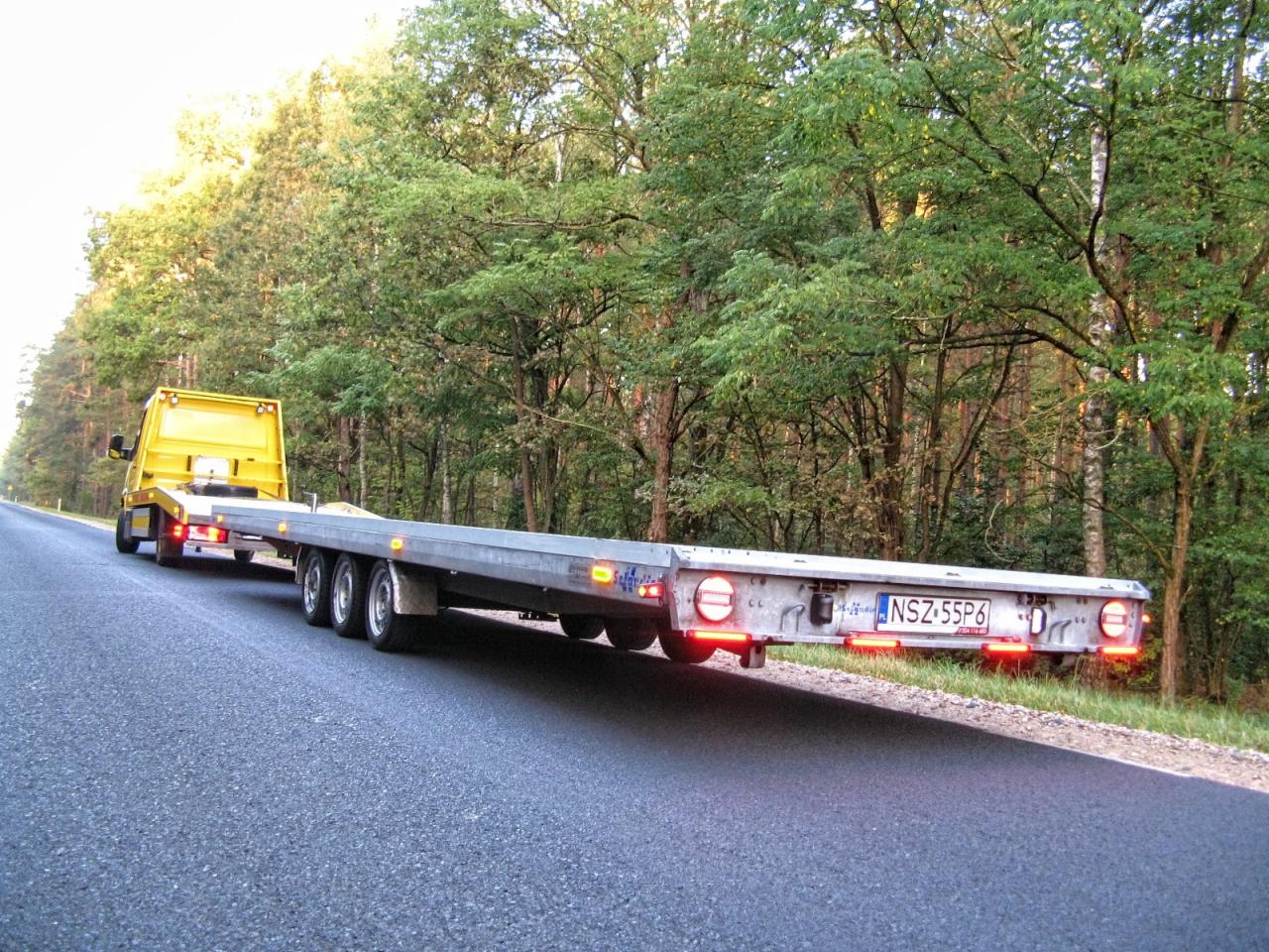 Żółta laweta z długą, szarą platformą na drodze asfaltowej, otoczona zielenią drzew. Widoczne światła stopu i tablica rejestracyjna NSZ 55P6.