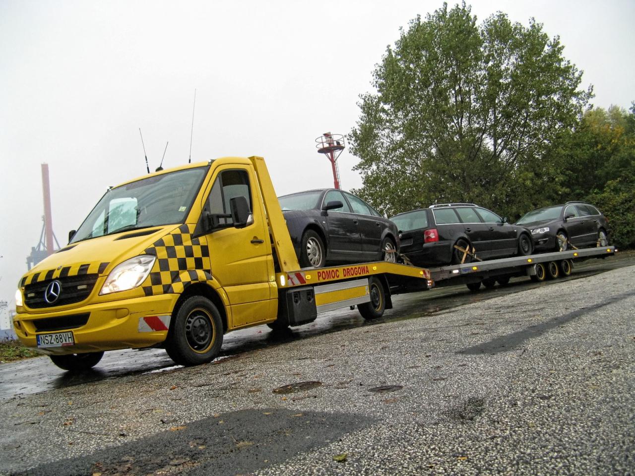 Żółta laweta Mercedes z szachownicą transportuje trzy czarne samochody osobowe na przyczepie. Widoczny napis 'Pomoc Drogowa'.