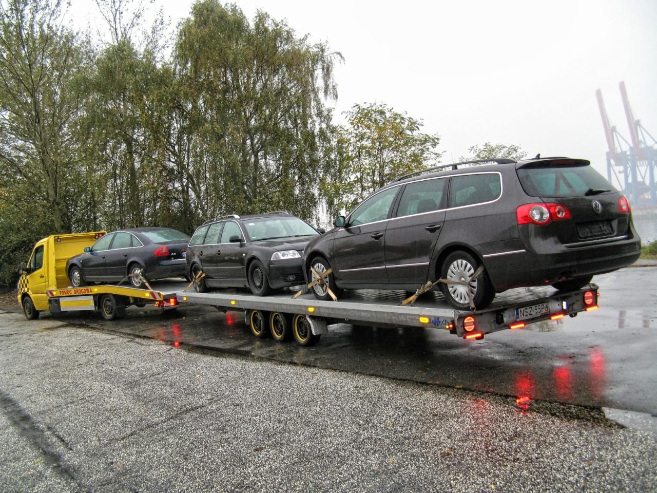 Żółta laweta z trzema samochodami osobowymi na platformie, transportowana w Rozogach. Widoczne drzewa w tle i dźwigi portowe w oddali.