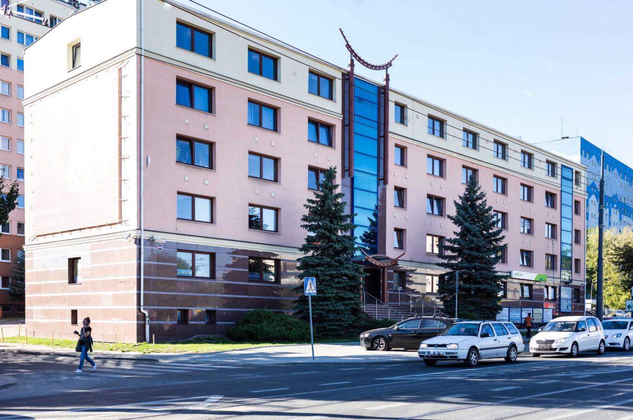 Czteropiętrowy budynek biurowy z przeszkloną klatką schodową, drzewami iglastymi przed wejściem i samochodami zaparkowanymi wzdłuż ulicy, widziany z perspektywy pieszego przechodzącego przez...