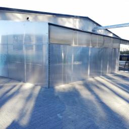 Arkanox - Zewnętrzna ściana hali produkcyjnej z przezroczystego poliwęglanu, z widocznymi stalowymi elementami konstrukcyjnymi i cieniem drzew na brukowanym chodniku.