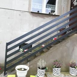 Arkanox - Zewnętrzna, metalowa balustrada schodowa w kolorze grafitowym, zamontowana przy wejściu do domu z surowym, betonowym wykończeniem, ozdobiona doniczkami z wrzosami i drobnymi dekoracjami.