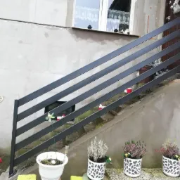 Zewnętrzna, metalowa balustrada schodowa w kolorze grafitowym, zamontowana przy wejściu do domu z surowym, betonowym wykończeniem, ozdobiona doniczkami z wrzosami i drobnymi dekoracjami.