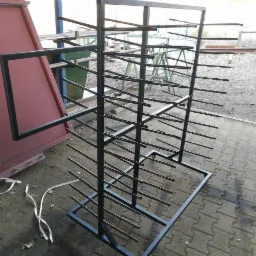 Stojak z czarnej stali z poziomymi prętami, widoczne elementy konstrukcyjne i otoczenie warsztatu, w tle konstrukcja pomalowana na czerwono.
