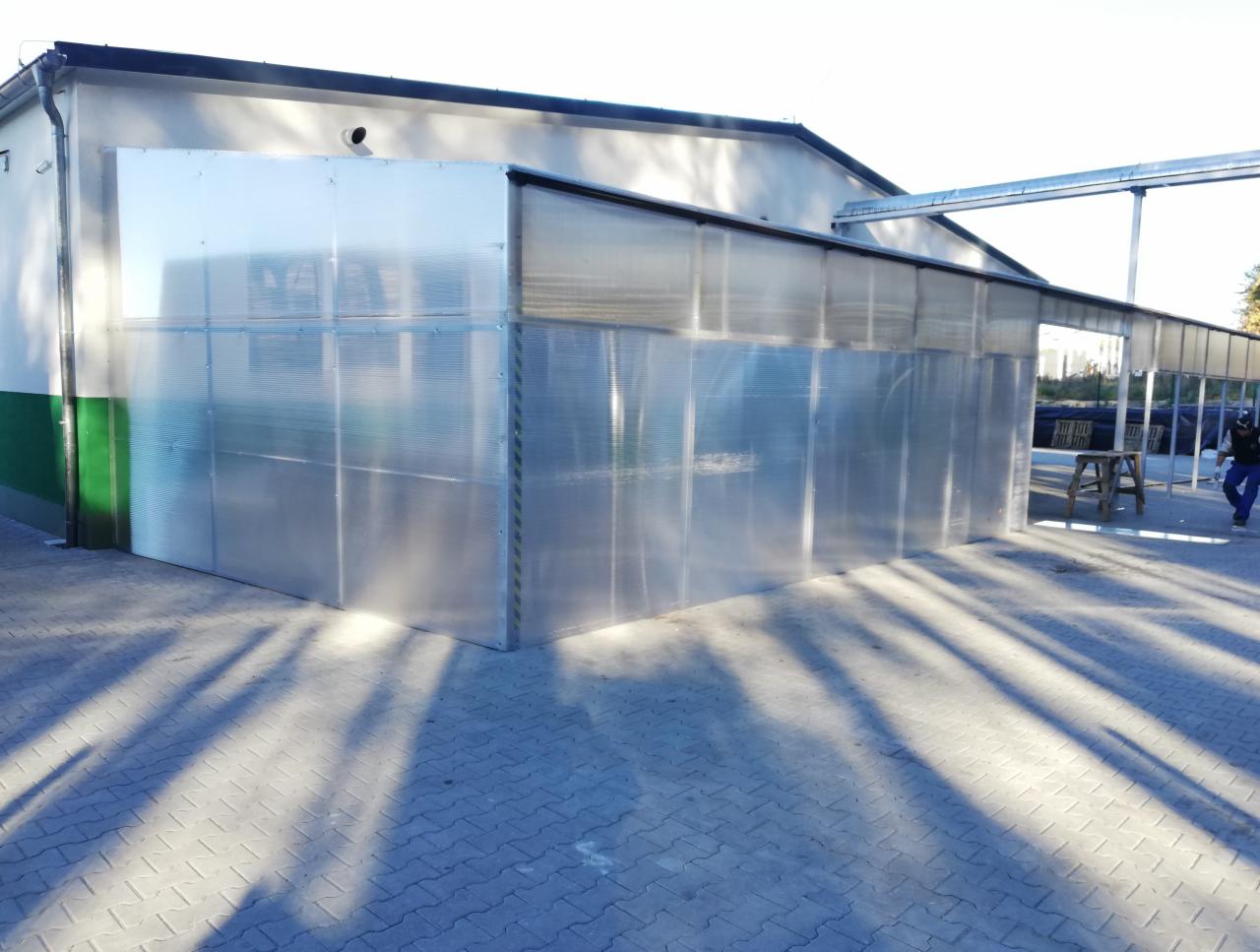 Zewnętrzna ściana hali produkcyjnej z przezroczystego poliwęglanu, z widocznymi stalowymi elementami konstrukcyjnymi i cieniem drzew na brukowanym chodniku.