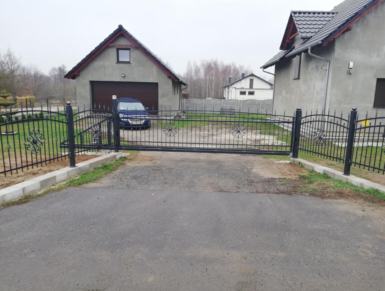 Czarna, metalowa brama wjazdowa z ozdobnymi elementami na posesji z domem i garażem w tle, widok z zewnątrz na szary, pochmurny dzień.