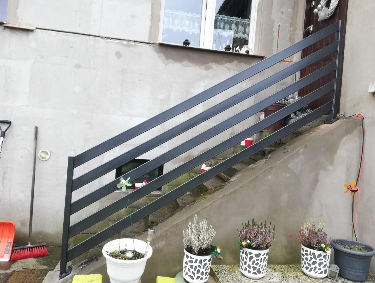 Zewnętrzna, metalowa balustrada schodowa w kolorze grafitowym, zamontowana przy wejściu do domu z surowym, betonowym wykończeniem, ozdobiona doniczkami z wrzosami i drobnymi dekoracjami.