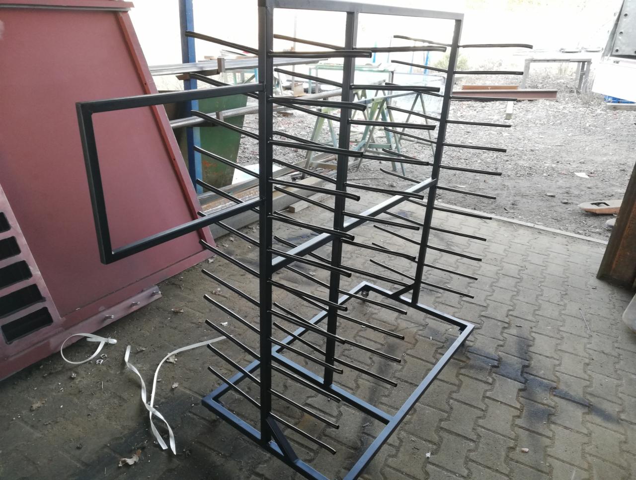 Stojak z czarnej stali z poziomymi prętami, widoczne elementy konstrukcyjne i otoczenie warsztatu, w tle konstrukcja pomalowana na czerwono.