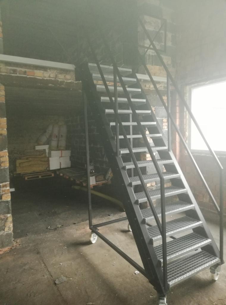 Przenośne schody stalowe z kratowymi stopniami i kółkami, umieszczone w surowym, ceglanym wnętrzu magazynu lub hali.