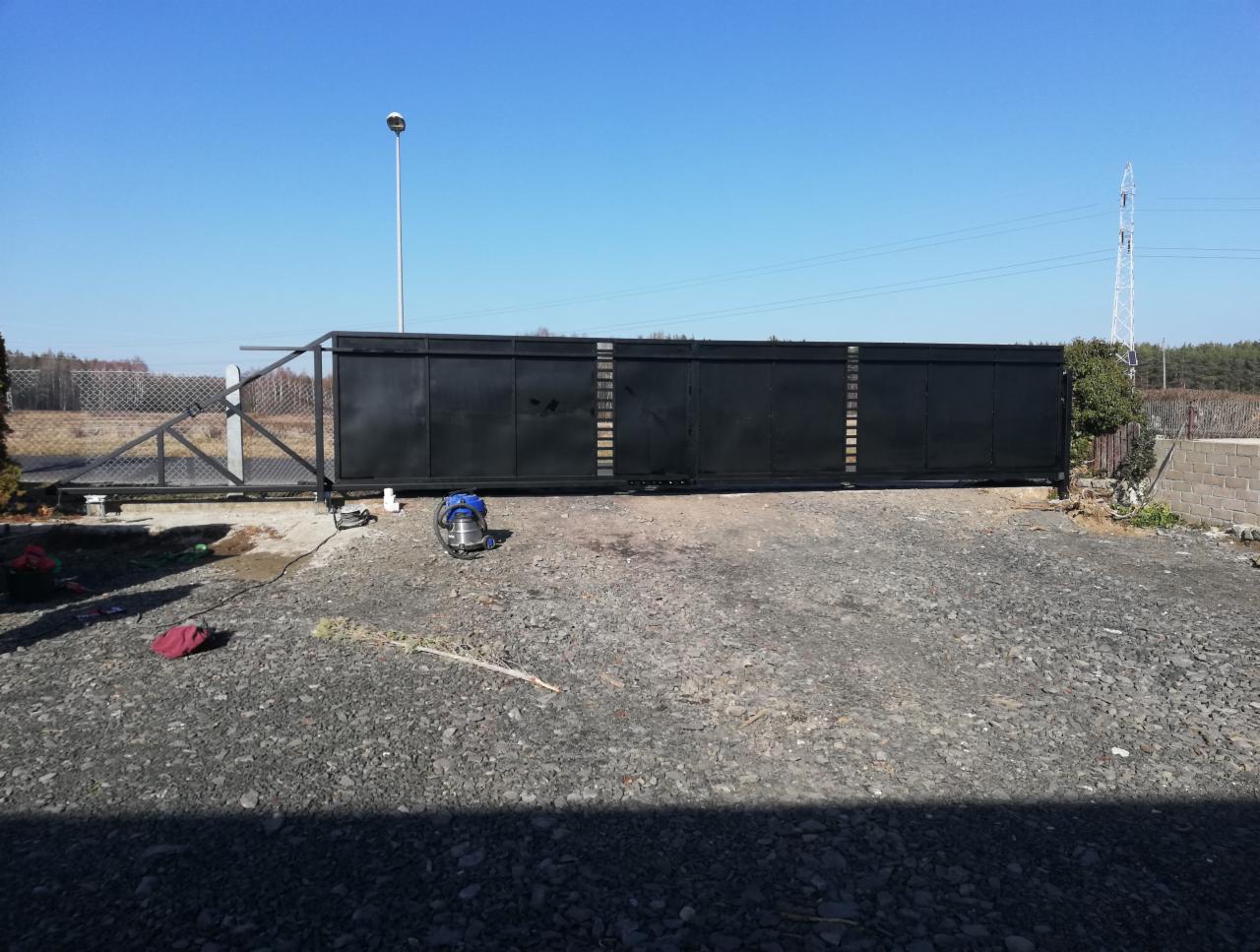 Czarna, przesuwna brama wjazdowa z metalową konstrukcją, widoczna na żwirowym podłożu w słoneczny dzień, częściowo zamontowana, ze sprzętem roboczym obok.