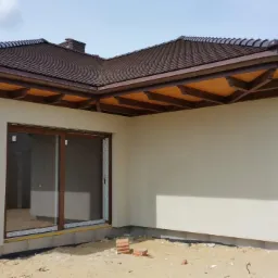 Wykończony budynek jednorodzinny z brązowym dachem i orynnowaniem, jasną elewacją i drewnianymi oknami, widoczne cegły na ziemi przed budynkiem.