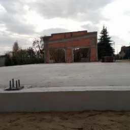 Surowy stan rozbudowy domu: wylana betonowa płyta fundamentowa z wystającymi śrubami kotwiącymi, częściowo wzniesione ściany z czerwonej cegły, widoczne drzewa w tle pod pochmurnym niebem.